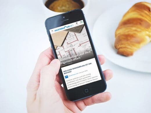Hand hält Smartphone mit Ratgeber-Artikel zum Thema Bodenbeläge. Auf dem Tisch ein Croissant und eine Tasse Kaffee.